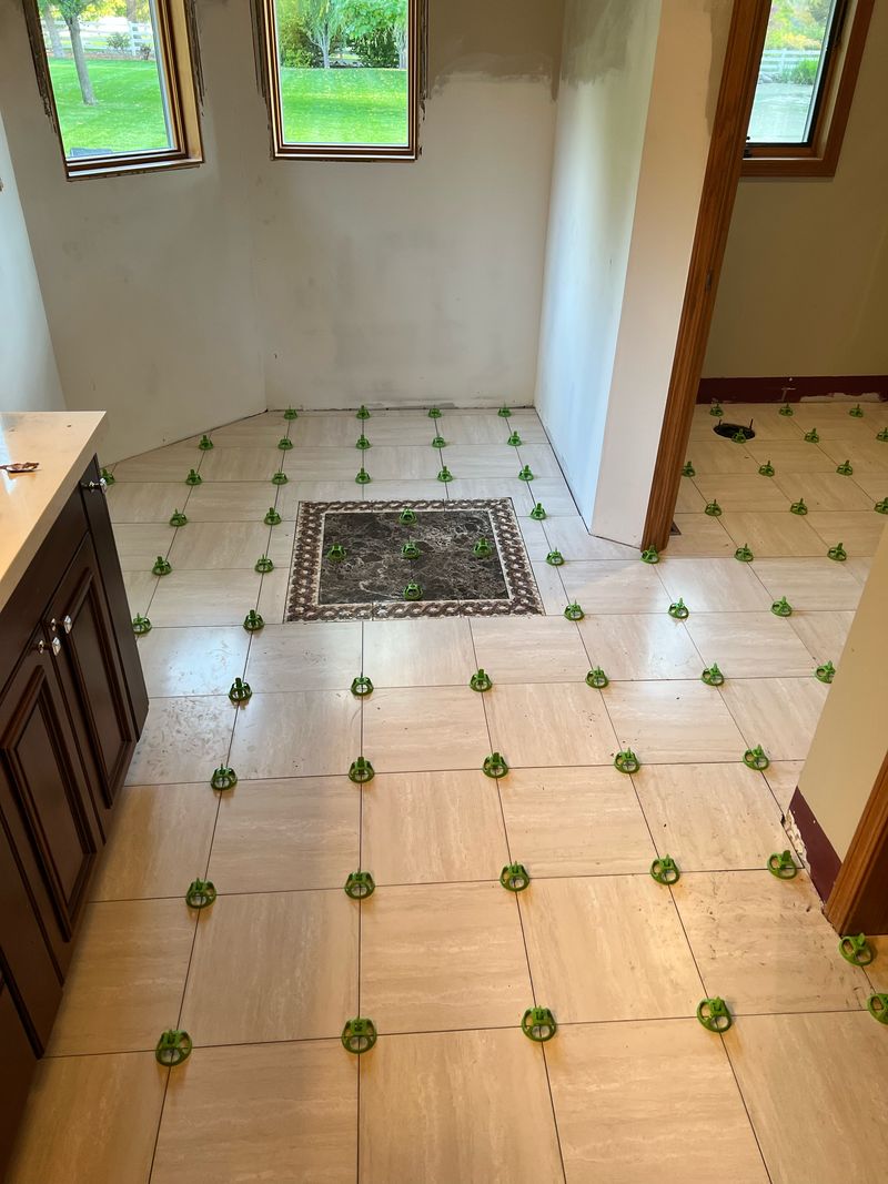 Large pale floor tile freshly set with green leveling clips around a dark stone inset