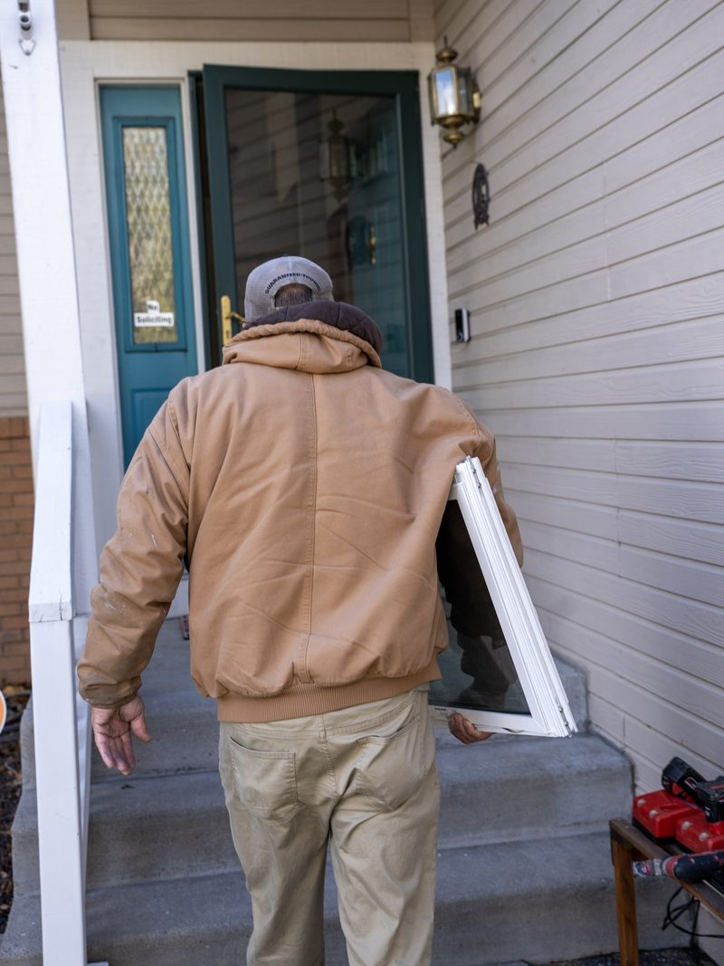 A worker in a tan jacket carrying a new white window unit up the front steps of a house