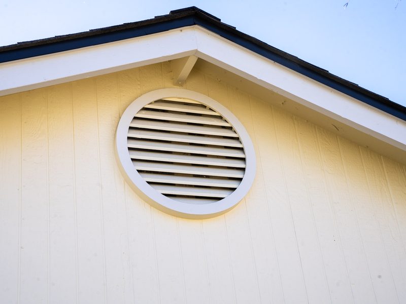Close view of a round louvred gable vent set high in the painted gable wall under a dark roof edge