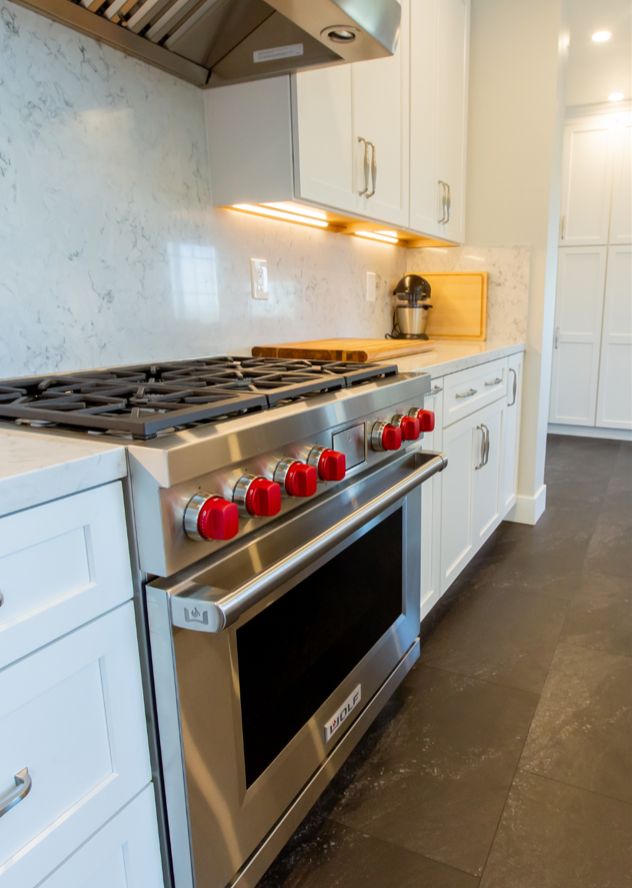 Professional-style gas range with red knobs under a stainless hood, against a stone-look backsplash with under-cabinet lights