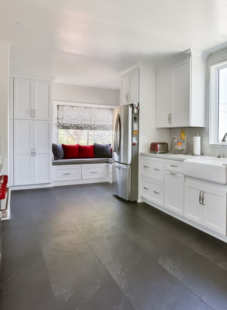 Remodeled kitchen with white shaker cabinets, a farmhouse sink, dark large-format floor tile and a cushioned window seat