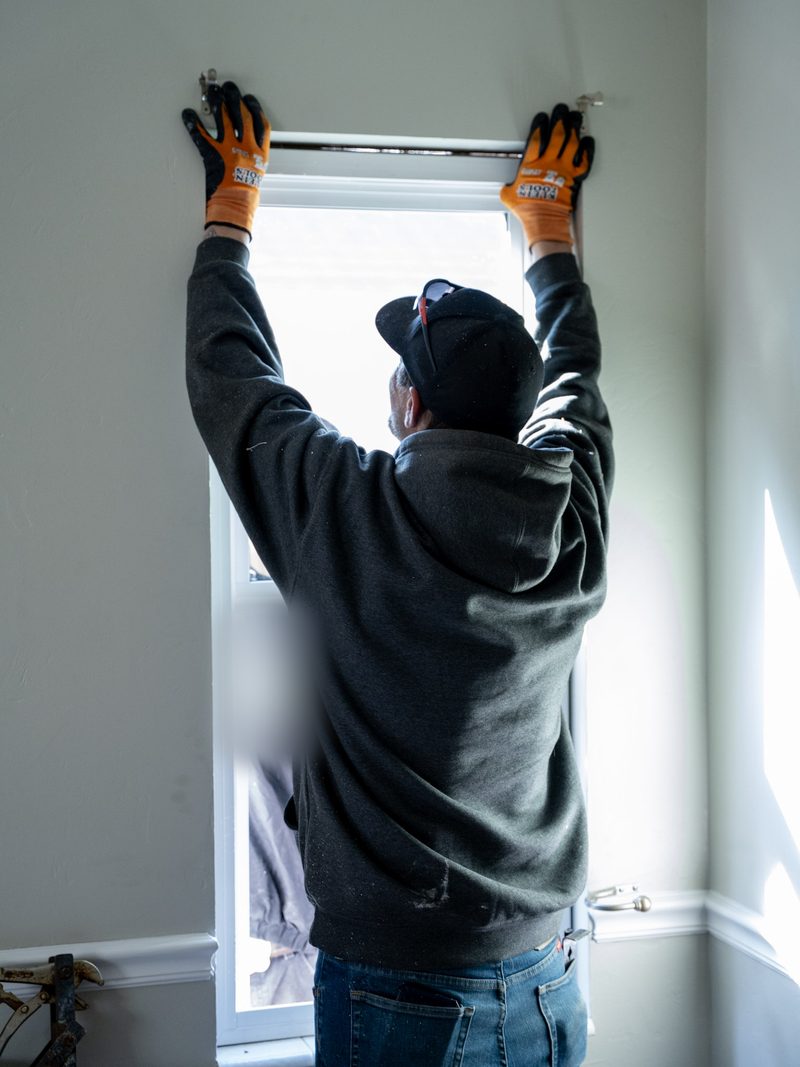 Seen from indoors, a worker in gloves holding a new window unit up against the head jamb of the opening