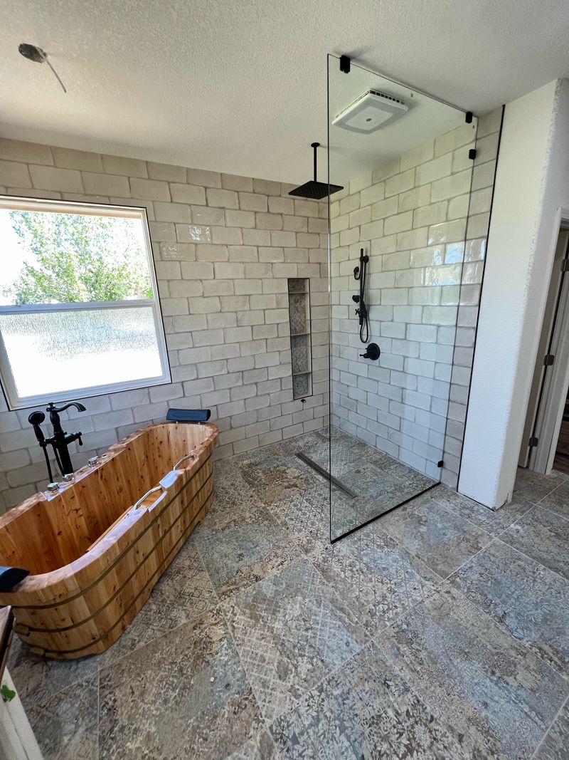 Walk-in shower with one glass panel, a ceiling rain head, a tiled niche and a linear drain beside a wooden soaking tub